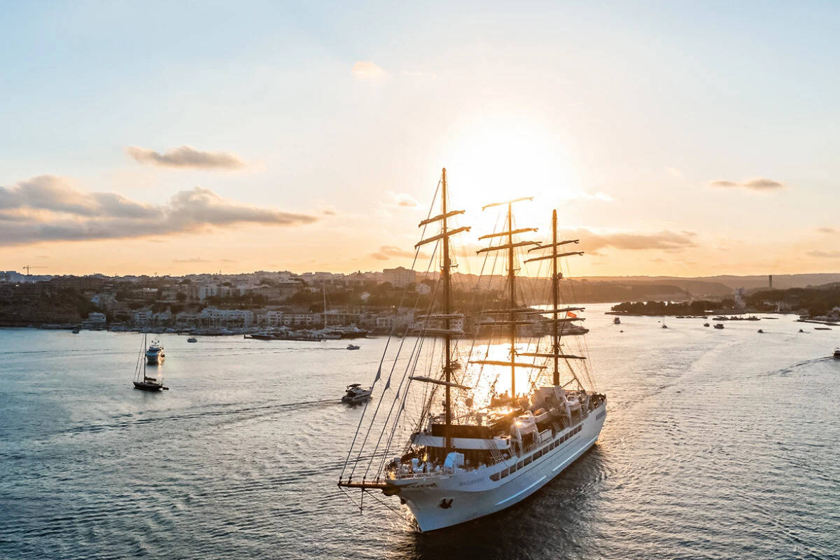 Ein Segelschiff mit drei Masten auf ruhigem Wasser bei Sonnenuntergang, im Hintergrund ist eine Küstenlinie mit Gebäuden und weiteren Booten.