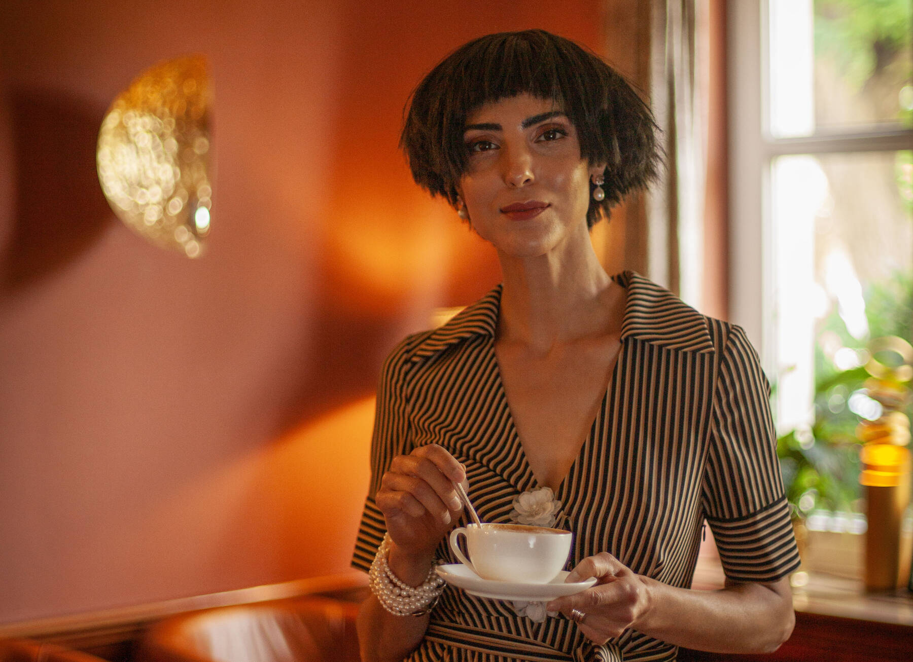 Eine Frau mit kurzem dunklem Haar und gestreiftem Kleid hält eine Kaffeetasse – warmes Licht und stilvolle Atmosphäre.
