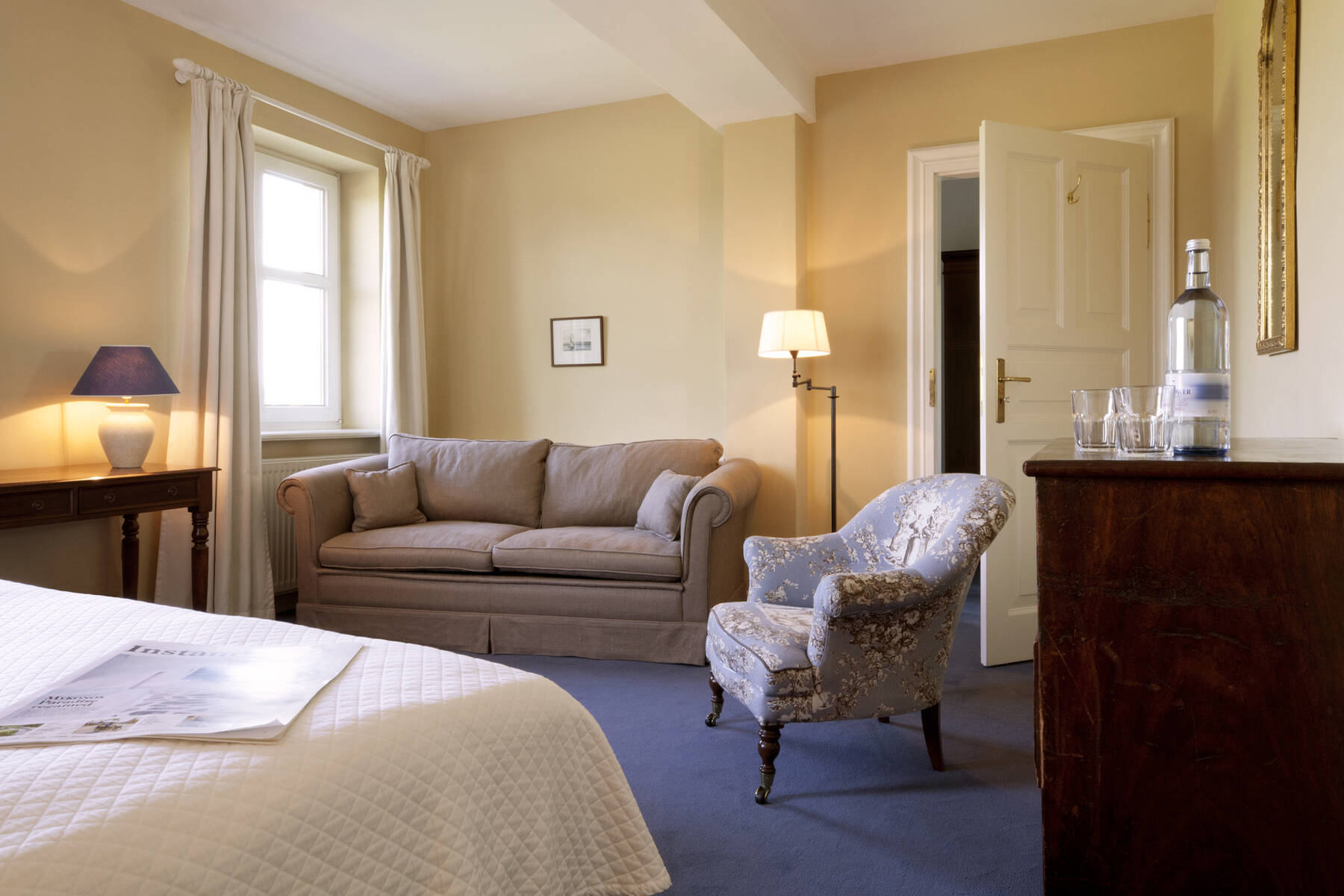 Ein elegantes Zimmer mit Bett, gemütlichem Sofa und Sessel – warme Farben und sanftes Licht schaffen Behaglichkeit.