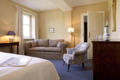 Ein elegantes Zimmer mit Bett, gemütlichem Sofa und Sessel – warme Farben und sanftes Licht schaffen Behaglichkeit.