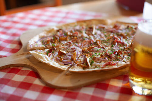 Ein knuspriger Flammkuchen liegt auf einem Holzbrett neben einem Glas Bier.