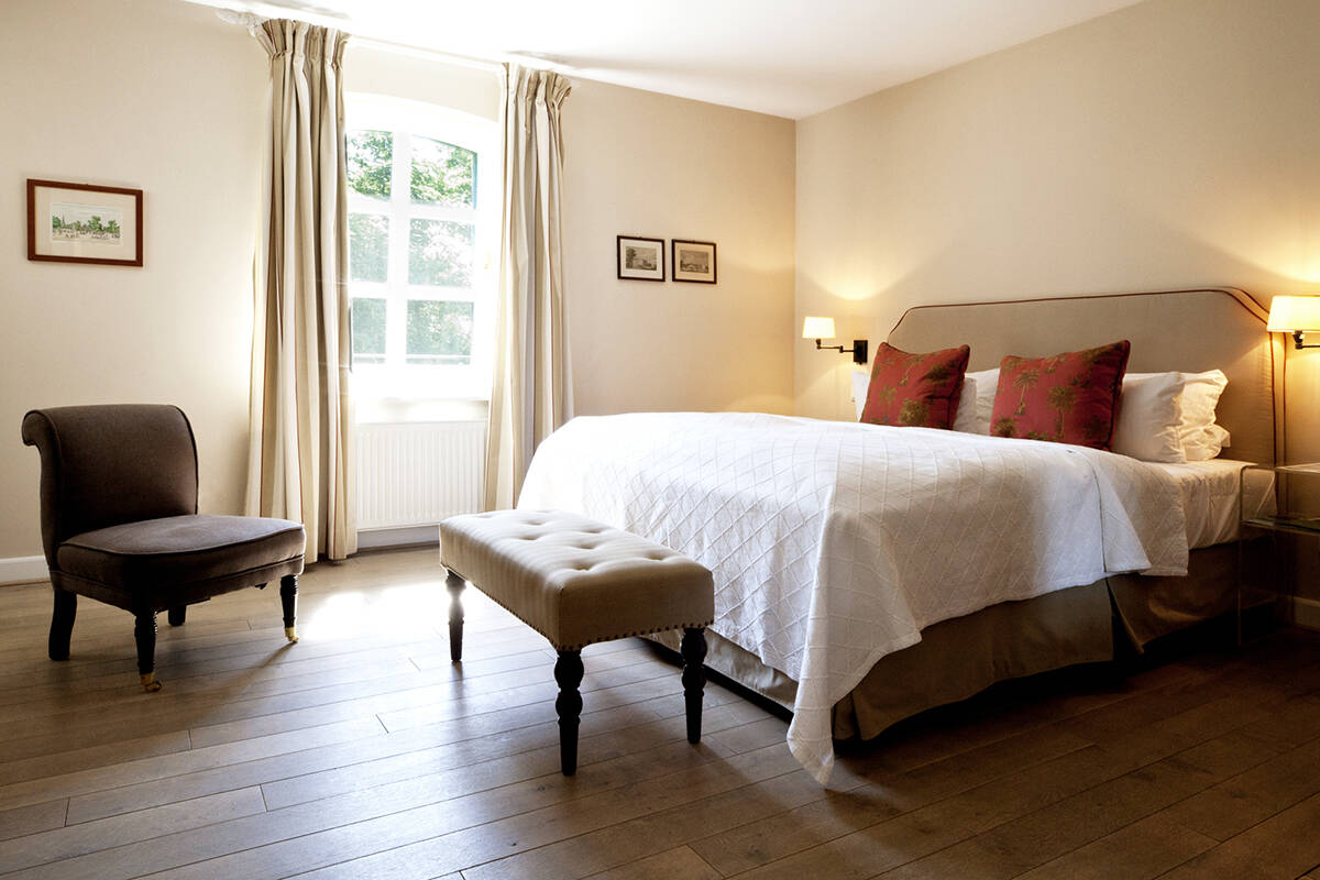 Ein hell eingerichtetes Schlafzimmer mit einem großen Bett, gepolsterten Hocker, Sessel, Vorhängen und drei gerahmten Bildern an der Wand.