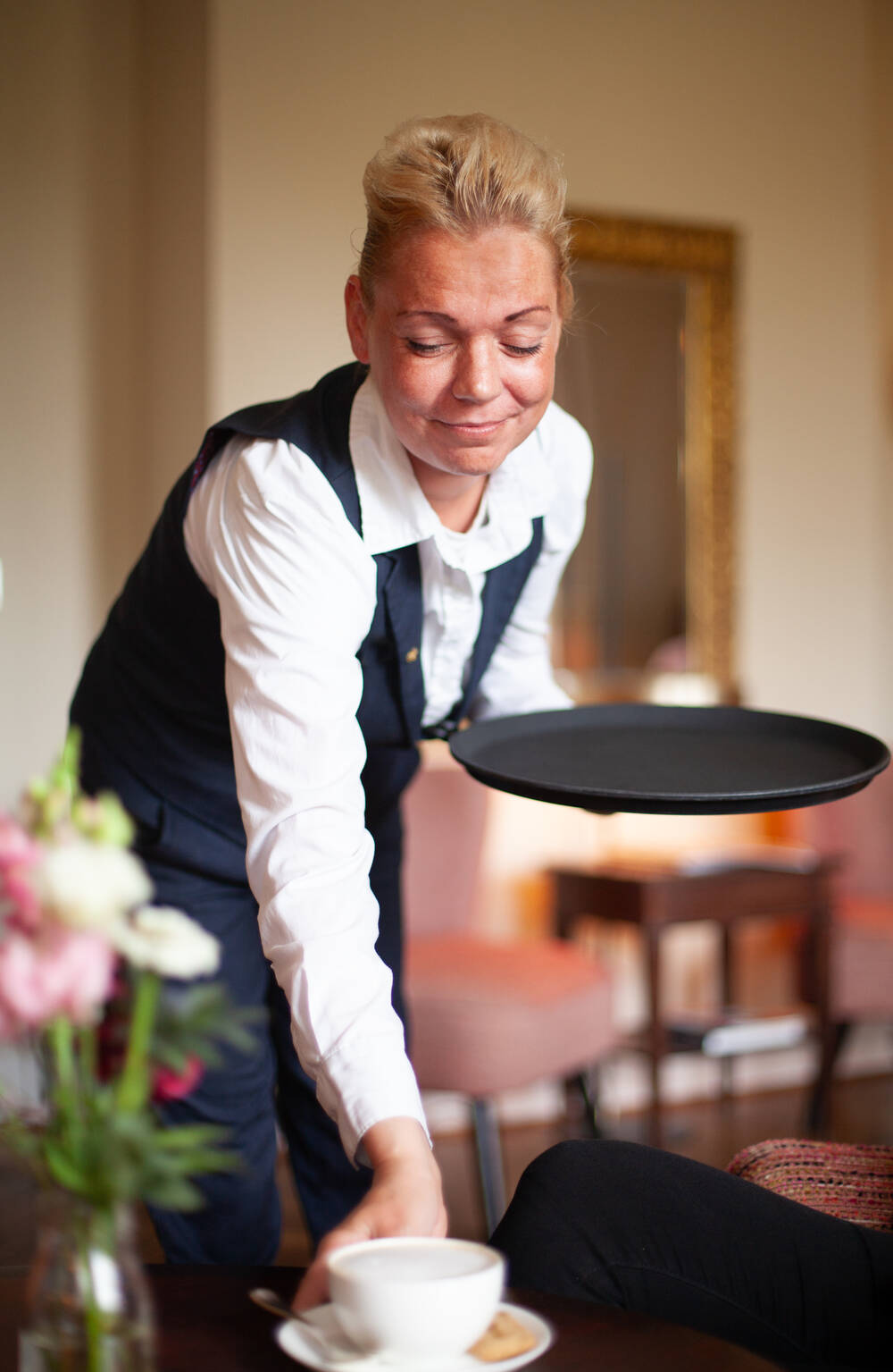 Eine Kellnerin in weißem Hemd und schwarzer Weste serviert eine Tasse Kaffee auf einem Tablett im Hotel Gutshaus Stolpe.