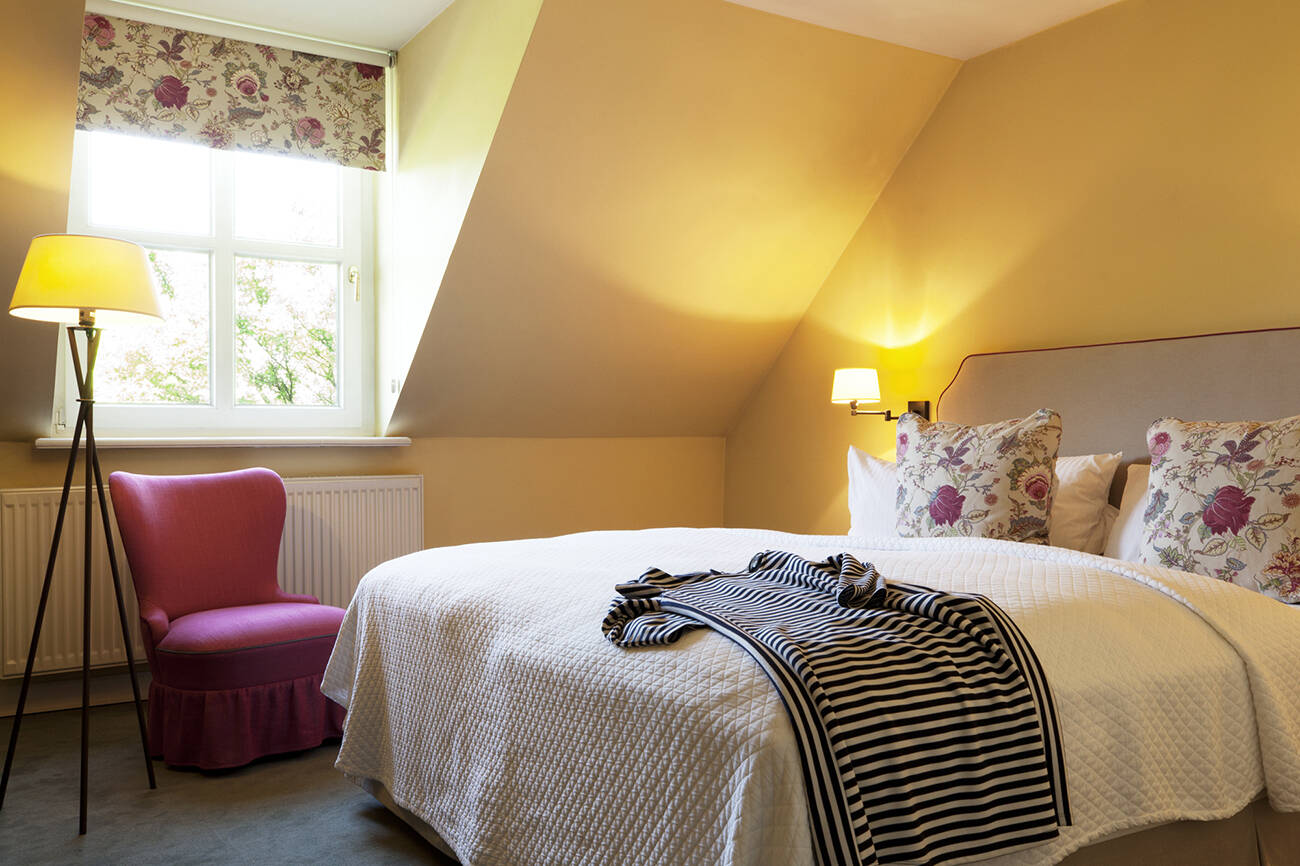 Ein Landhauszimmer mit einem weißen Bett, gestreiften Pullover darauf, rosa Sessel, einer Stehlampe und einem Fenster mit Rollo.