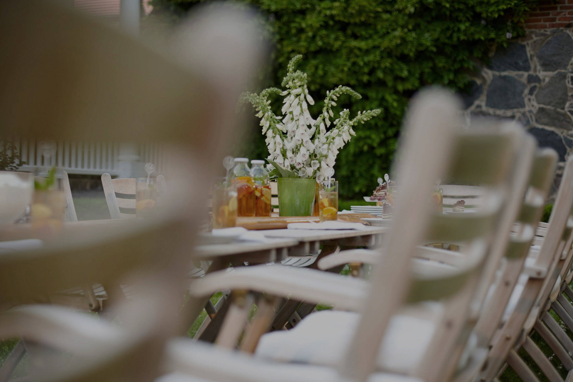 Ein festlich gedeckter Gartentisch mit weißen Blüten, Getränken und Holzstühlen in ruhiger, grüner Umgebung.