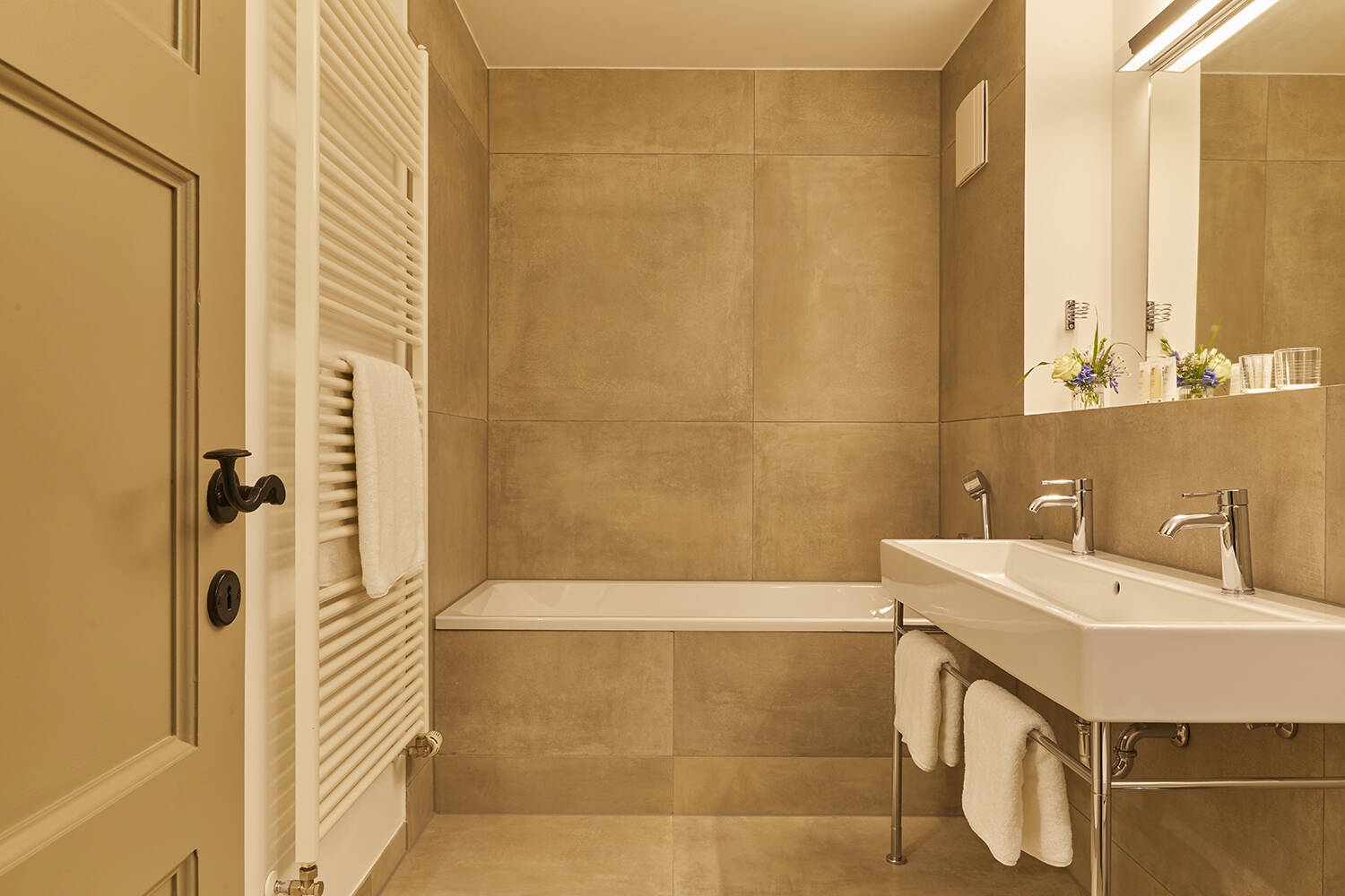 Ein modernes Badezimmer mit einer Badewanne, einem Doppelwaschbecken, großen Spiegel und hellen Fliesen in warmen Beigetönen.