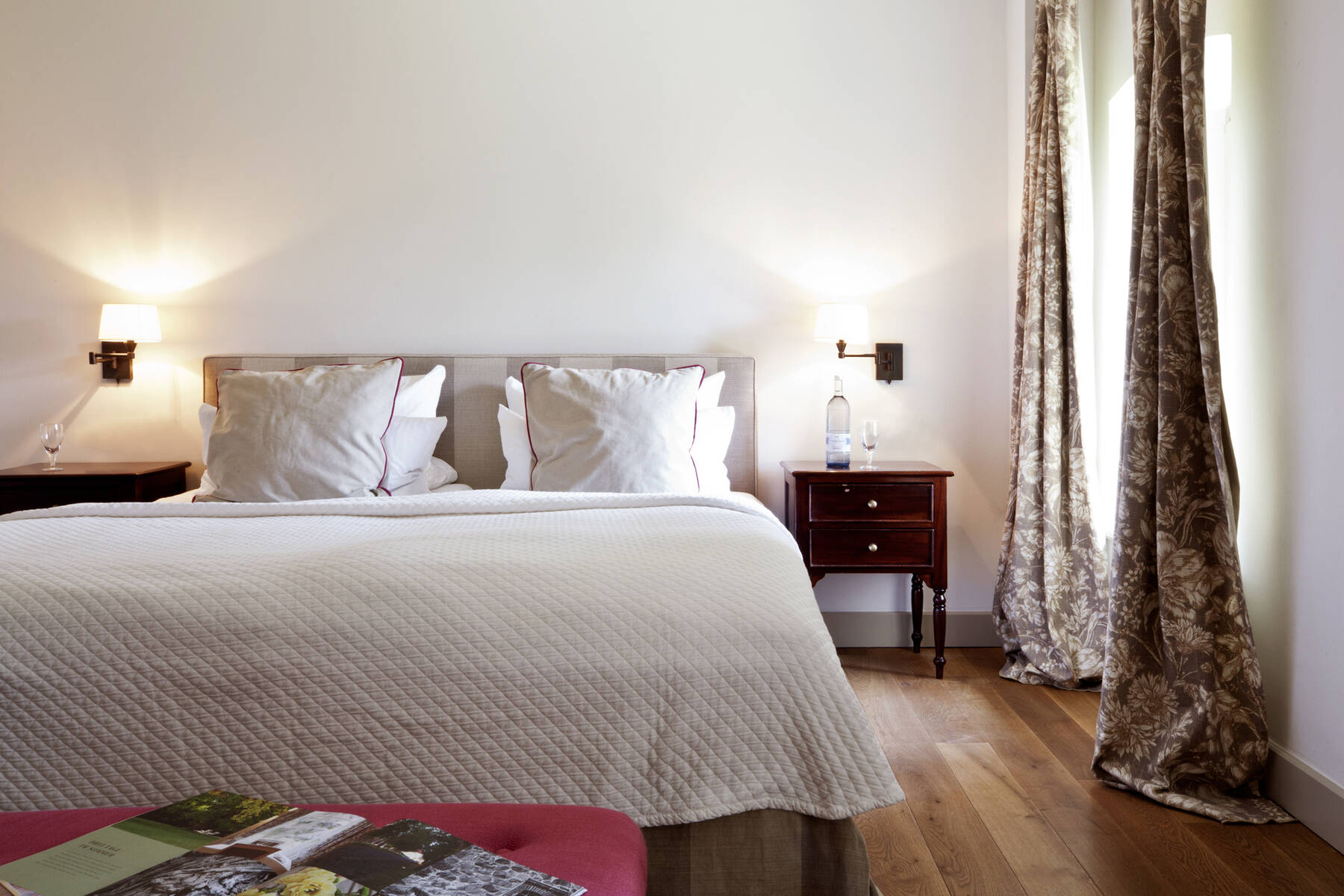 Ein helles Hotelzimmer mit einem großen Doppelbett, weißen Kissen, Nachttischen, Lampen und Vorhängen in Naturtönen.