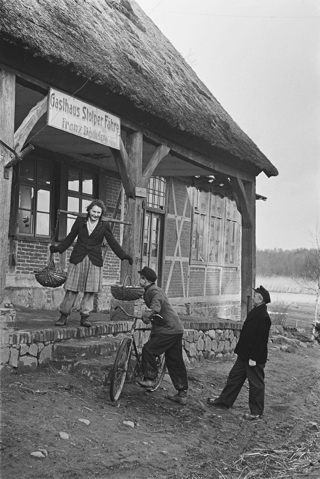 Ein Schwarz-weißes Foto vor dem 'Gasthaus Stolper Fähre' mit drei Personen, darunter eine Frau mit Körben und ein Mann mit Fahrrad.