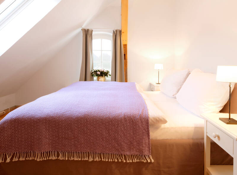 Eine Hotel Suite mit einem Bett mit einer lila Decke darauf, zwei Nachttischen mit Lampen und einem Fenster mit Vorhängen im Hintergrund.
