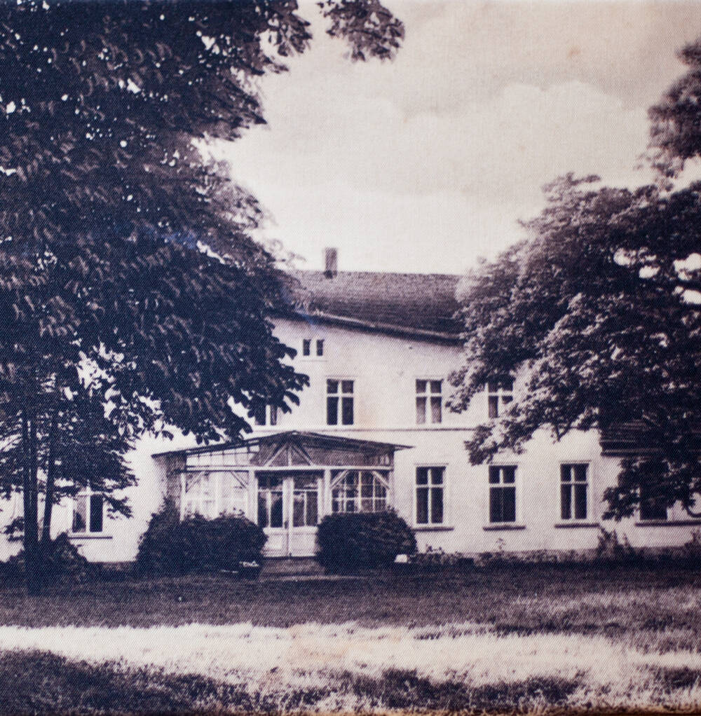 Ein zweistöckiges Gebäude mit einer Veranda, umgeben von großen Bäumen und einer Rasenfläche im Vordergrund, dargestellt als ein altes schwarz-weißes Bild.