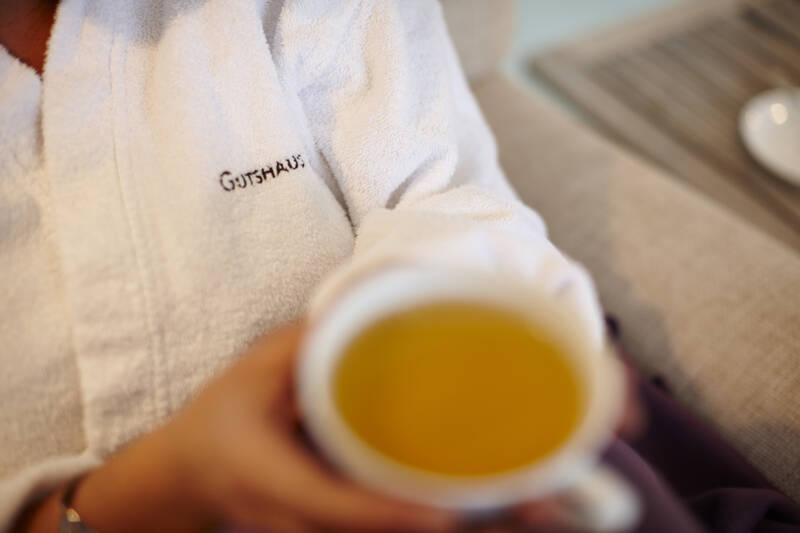 Eine Person in einem weißen Bademantel mit der Aufschrift „Gutshaus“ hält eine Tasse heißen Tee – Entspannung pur.