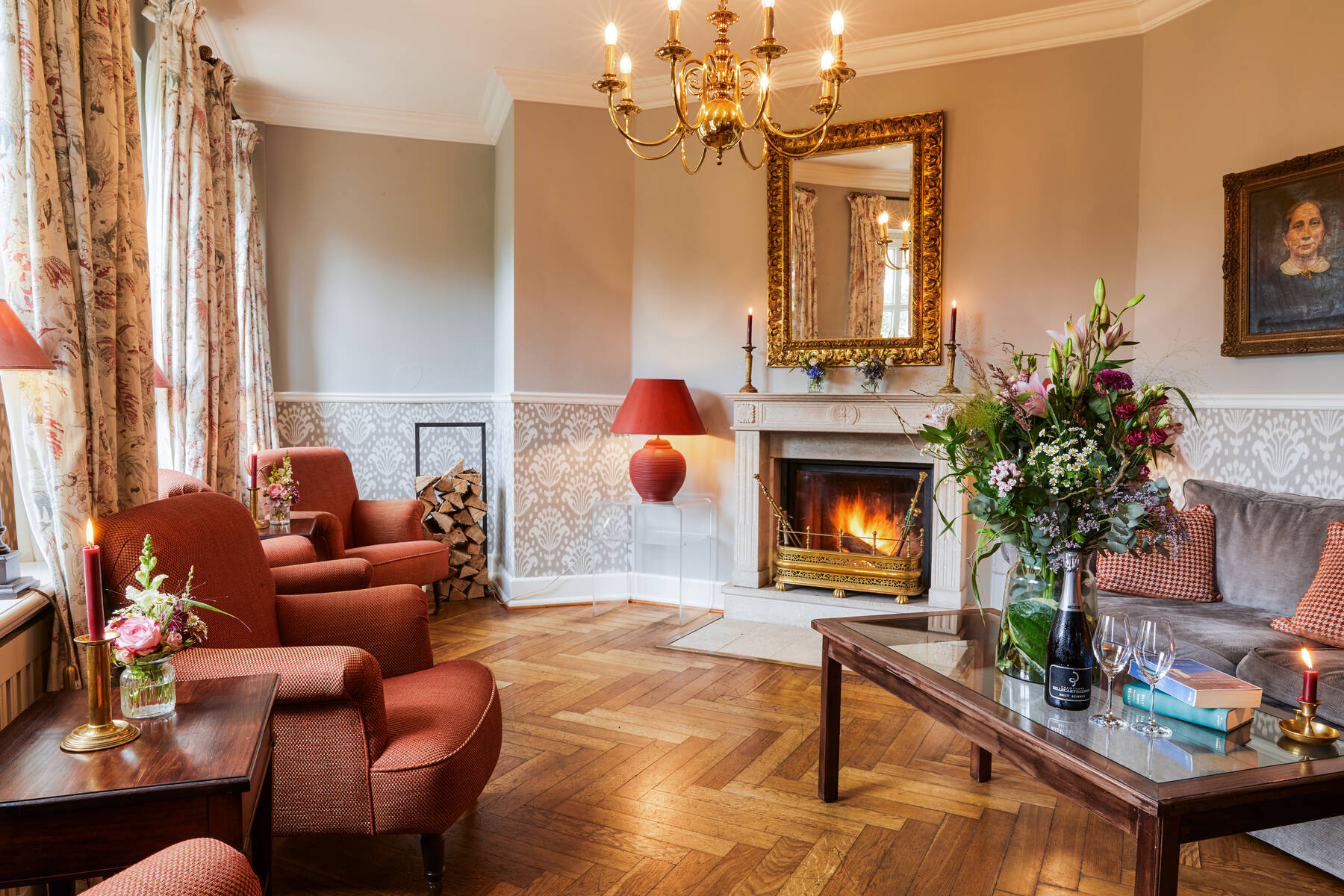 Eine elegante Hotel Suite mit roten Sesseln, einem Kamin, Blumenstrauß und goldenen Spiegel schafft eine warme, gemütliche Atmosphäre.