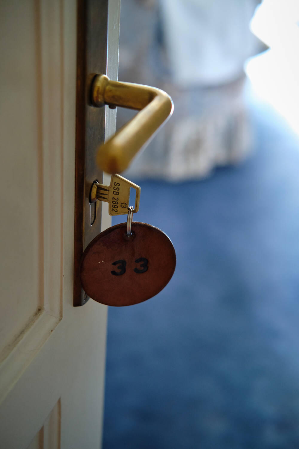 Ein goldener Türgriff mit einem Zimmerschlüssel und rundem Anhänger, dahinter ein Hotelzimmer mit blauem Teppich.