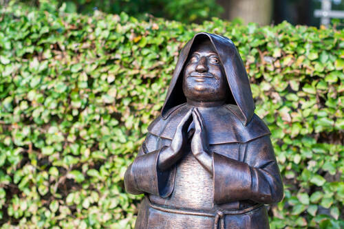 Eine bronzene Statue eines Mönchs mit Kapuze und gefalteten Händen vor einer grünen Hecke im Garten des Gutshauses Stolpe.