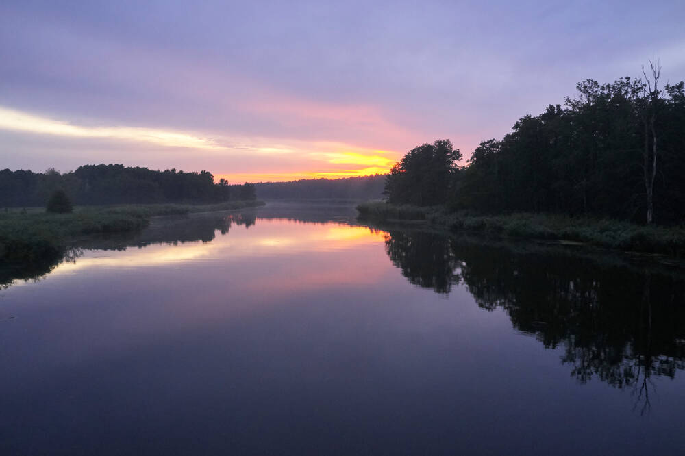Ein ruhiger Fluss im Morgenlicht, der Himmel leuchtet in Rosa und Violett – friedvolle Naturstimmung.