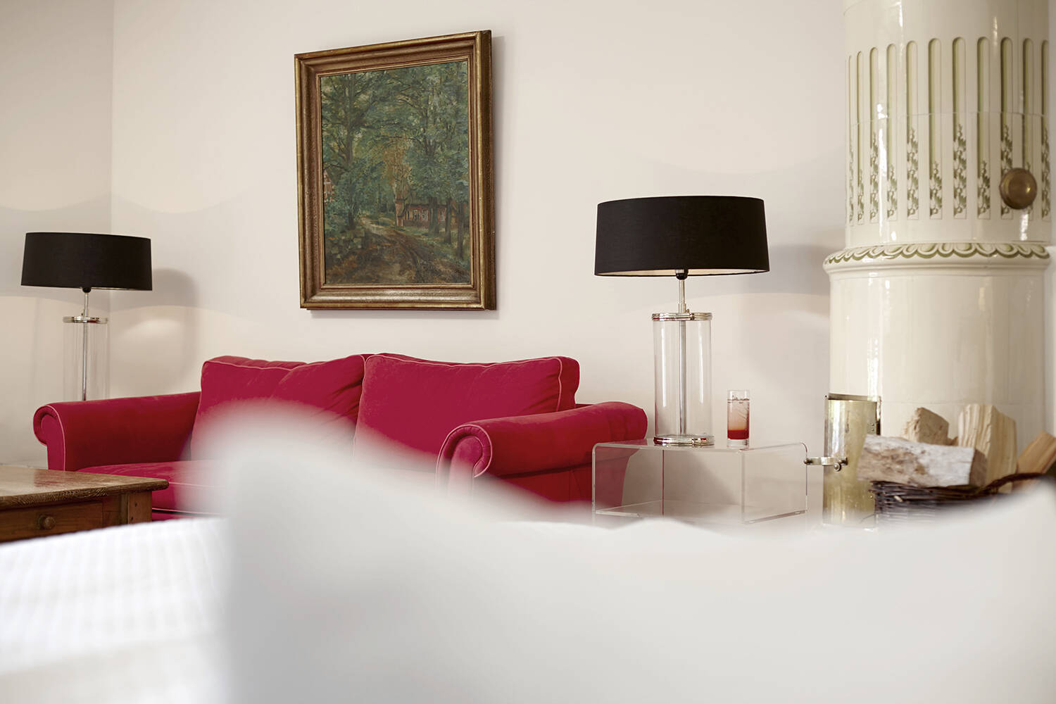 Ein rotes Sofa mit zwei schwarzen Lampen auf transparenten Beistelltischen, ein Gemälde an der Wand darüber und ein weißer Kachelofen rechts im Raum.