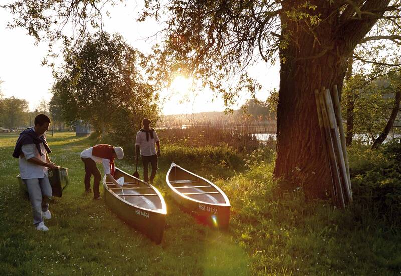 Drei Personen bereiten bei Sonnenuntergang zwei Kanus am Flussufer für eine Fahrt vor.