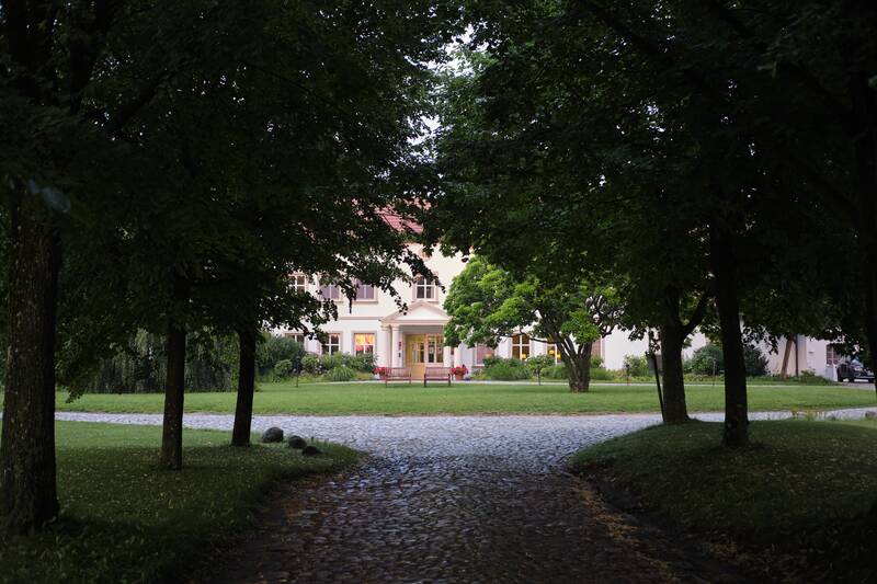 Ein Kopfsteinpflasterweg zwischen Bäumen führt zu einem hellen zweistöckigen Haus mit Säuleneingang.