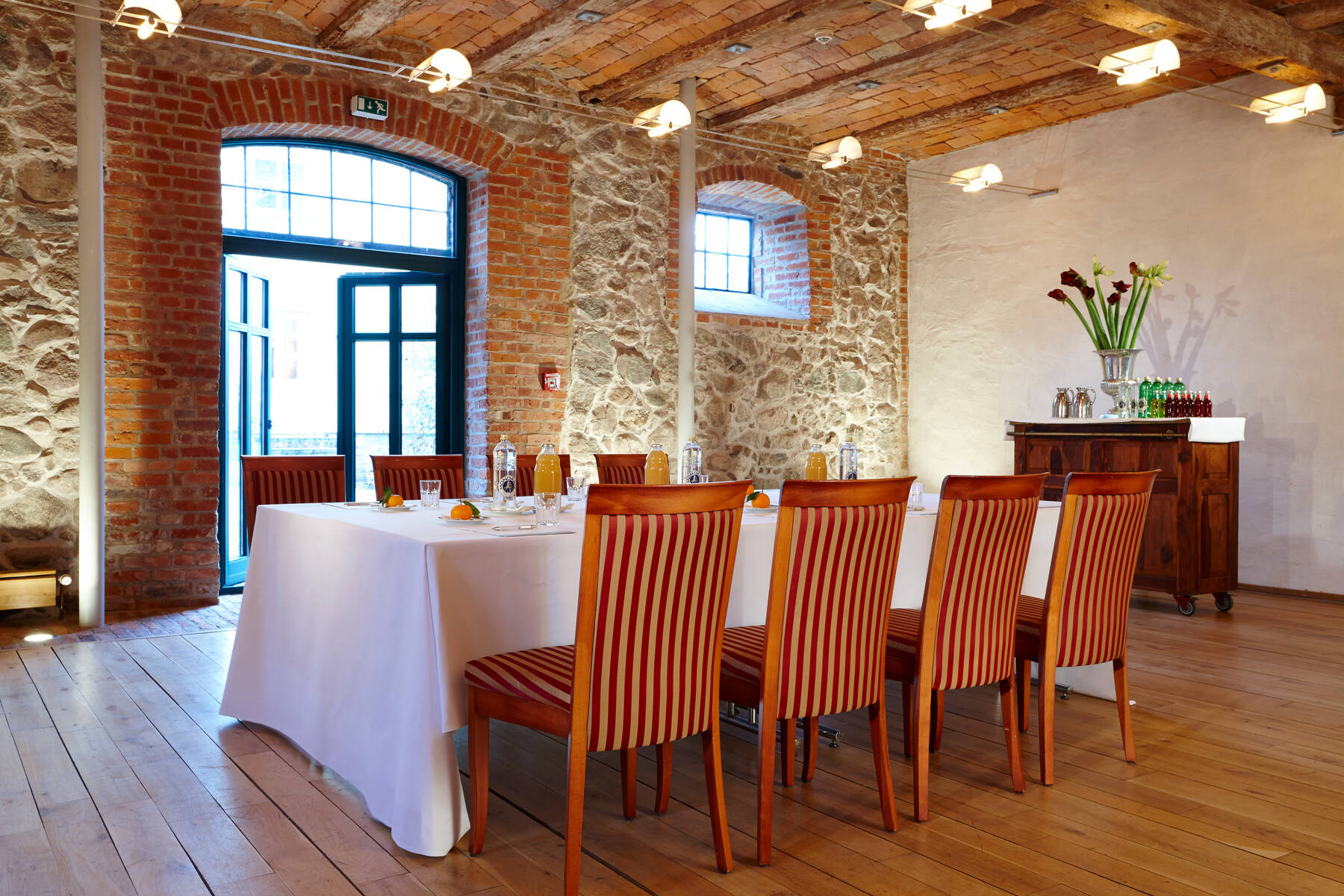 Ein Tagungsraum nahe Anklam mit einem Holzboden, Steinwänden und einem Tisch mit weißer Tischdecke, umgeben von Holzstühlen mit rot-weiß gestreiftem Bezug.
