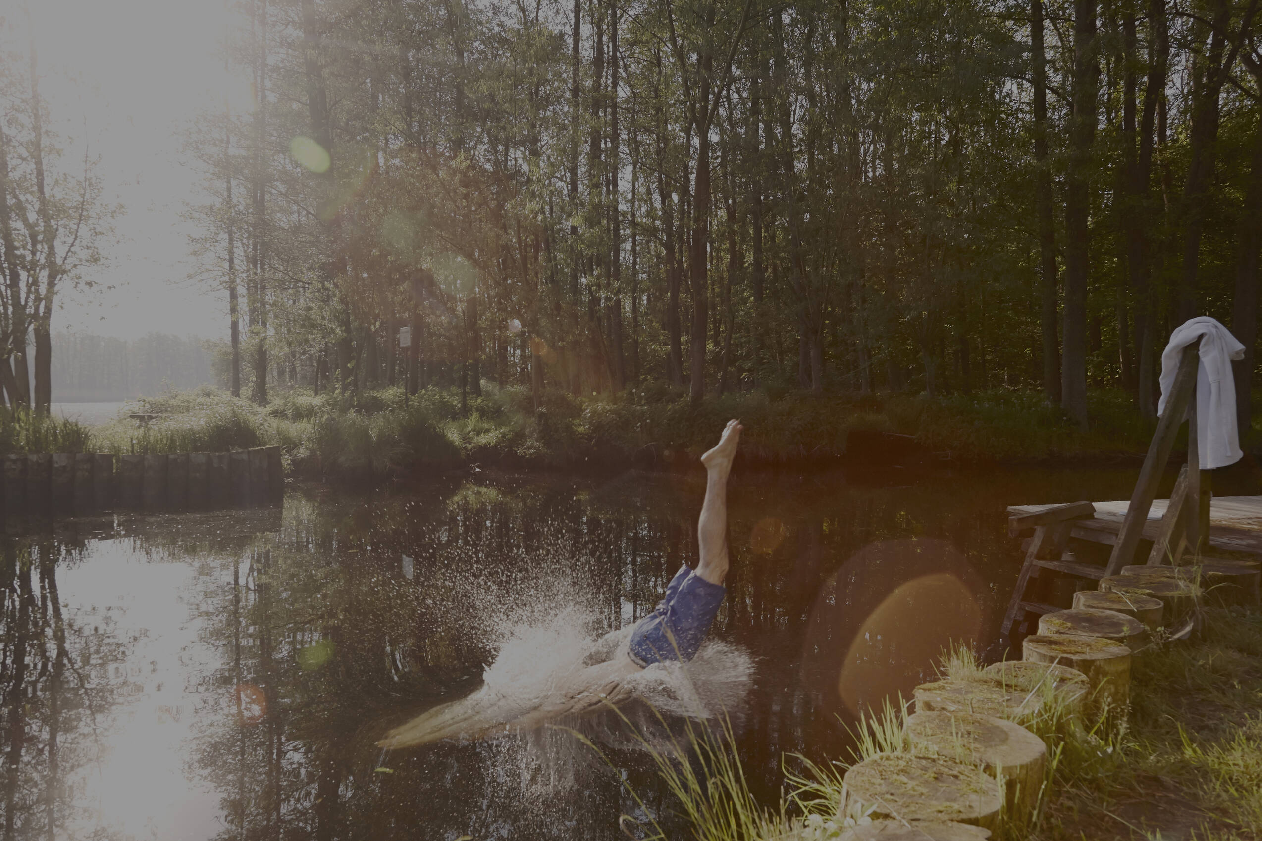 Eine Person taucht in einen See ein, Spritzwasser in der Sonne, Wald und Steg im Hintergrund.