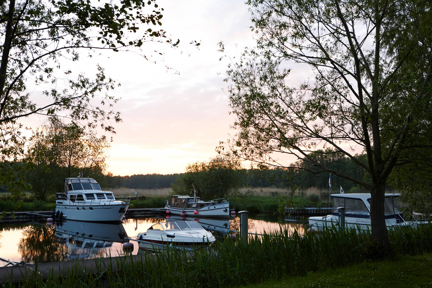 Mehrere Boote liegen still im kleinen Hafen, umgeben von Bäumen, während die Sonne sanft hinter dem Wald untergeht.