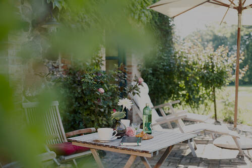 Ein Holztisch mit Getränken und Blumen steht auf einer sonnigen Terrasse mit Blick auf den Garten und eine Liege.