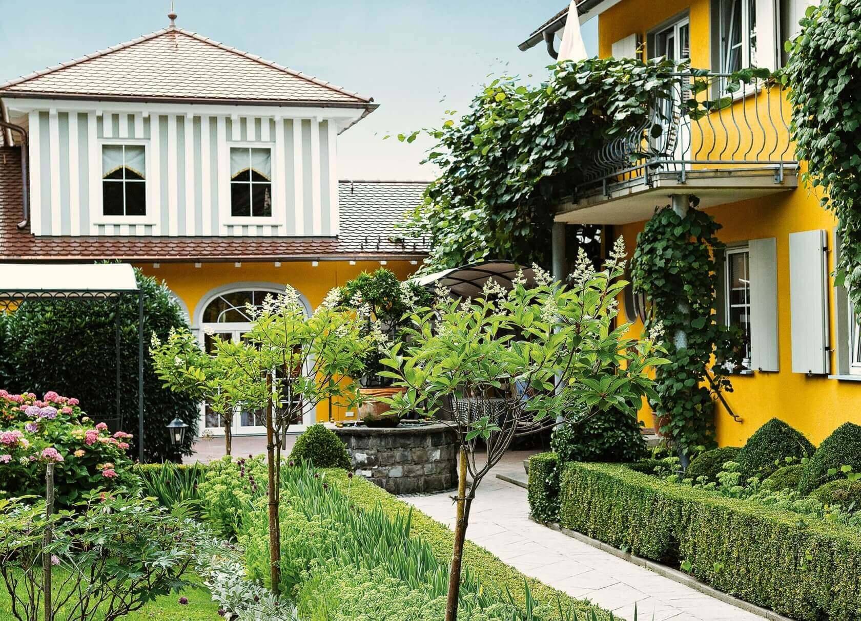 Ein sonniger Garten mit gepflegten Pflanzen und gelbem Gebäude mit Balkonen und weißen Fensterläden.