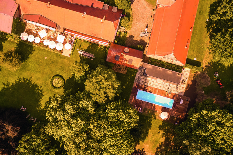 Eine Luftaufnahme zeigt ein Anwesen mit roten Dächern, gepflegtem Garten und blauem Pool zwischen alten Bäumen.