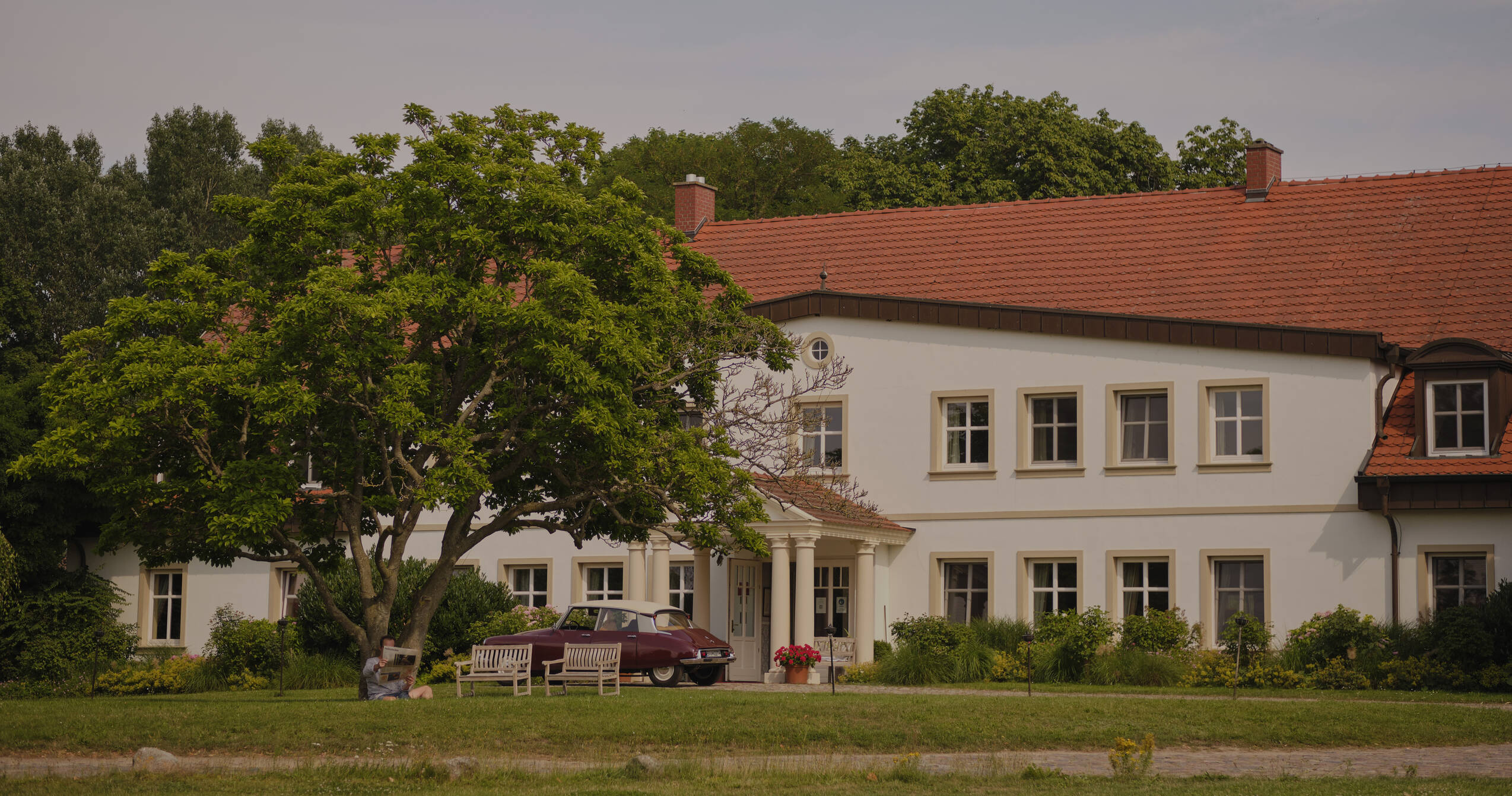 Ein elegantes Gutshaus mit einem roten Dach und weißen Säulen, umgeben von grünen Bäumen und einem alten Auto im Vordergrund.