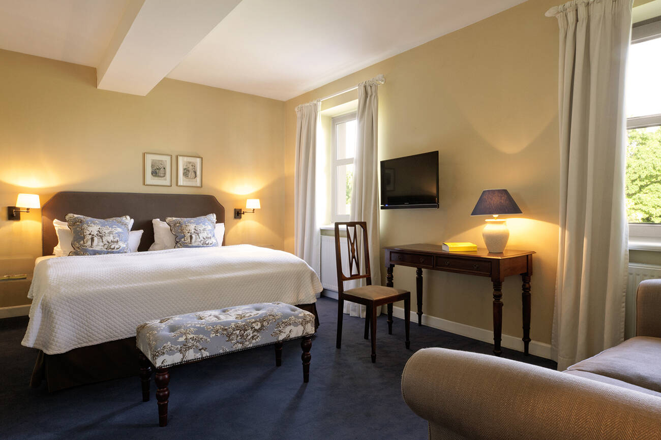 Ein helles Hotelzimmer mit einem Doppelbett, Schreibtisch, Fernseher, Sessel und Fenster mit weißen Vorhängen.