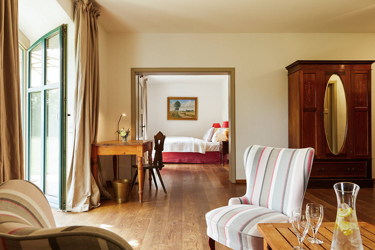Ein helles elegantes Hotelzimmer mit einem Holzboden, Fenstern mit Vorhängen, einem Schreibtisch, Stuhl, Zugang zum Balkon und einem angrenzenden Schlafzimmer.
