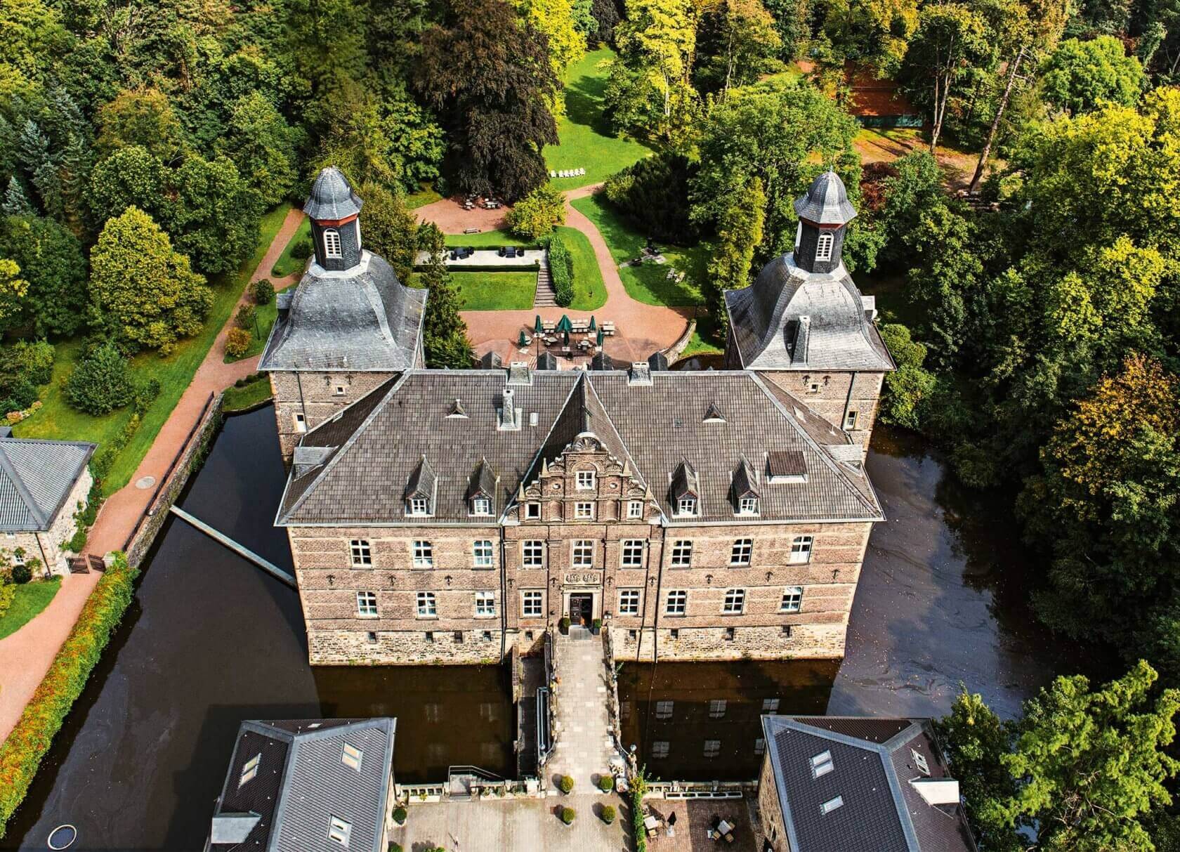 Ein historisches Wasserschloss mit Türmen und Innenhof, umgeben von Wassergräben und einer grünen Parklandschaft.