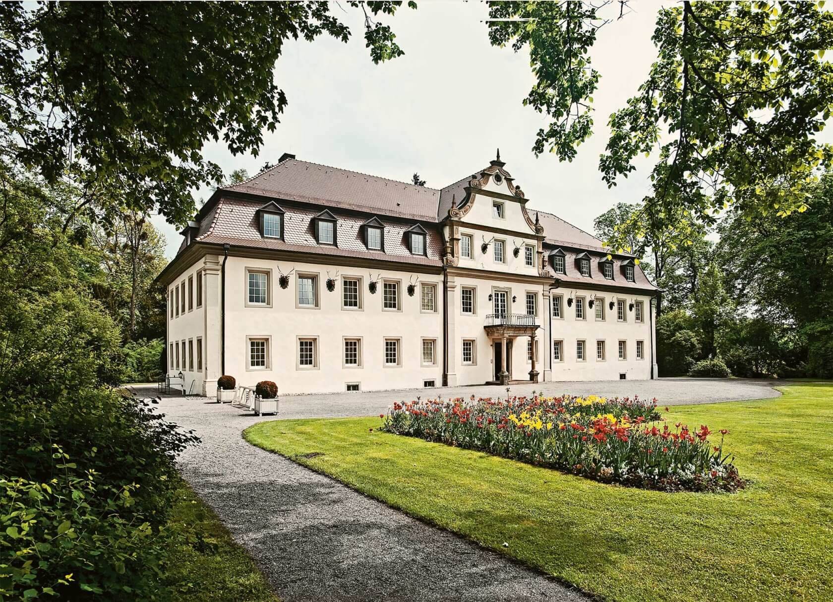 Ein barockes Herrenhaus mit einem Satteldach, mehreren Dachgauben und einer symmetrischen Fensteranordnung, umgeben von Bäumen und einem Blumenbeet.