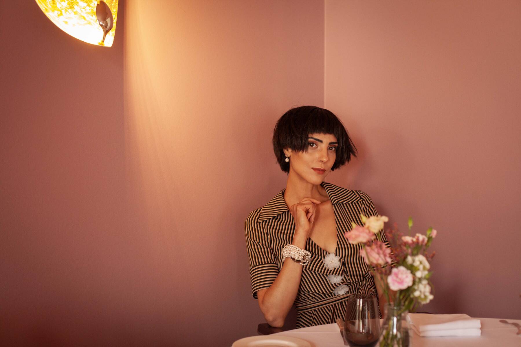 Eine Frau mit kurzem, dunklem Haar sitzt an einem Tisch mit Blumen und blickt ruhig in die Kamera.