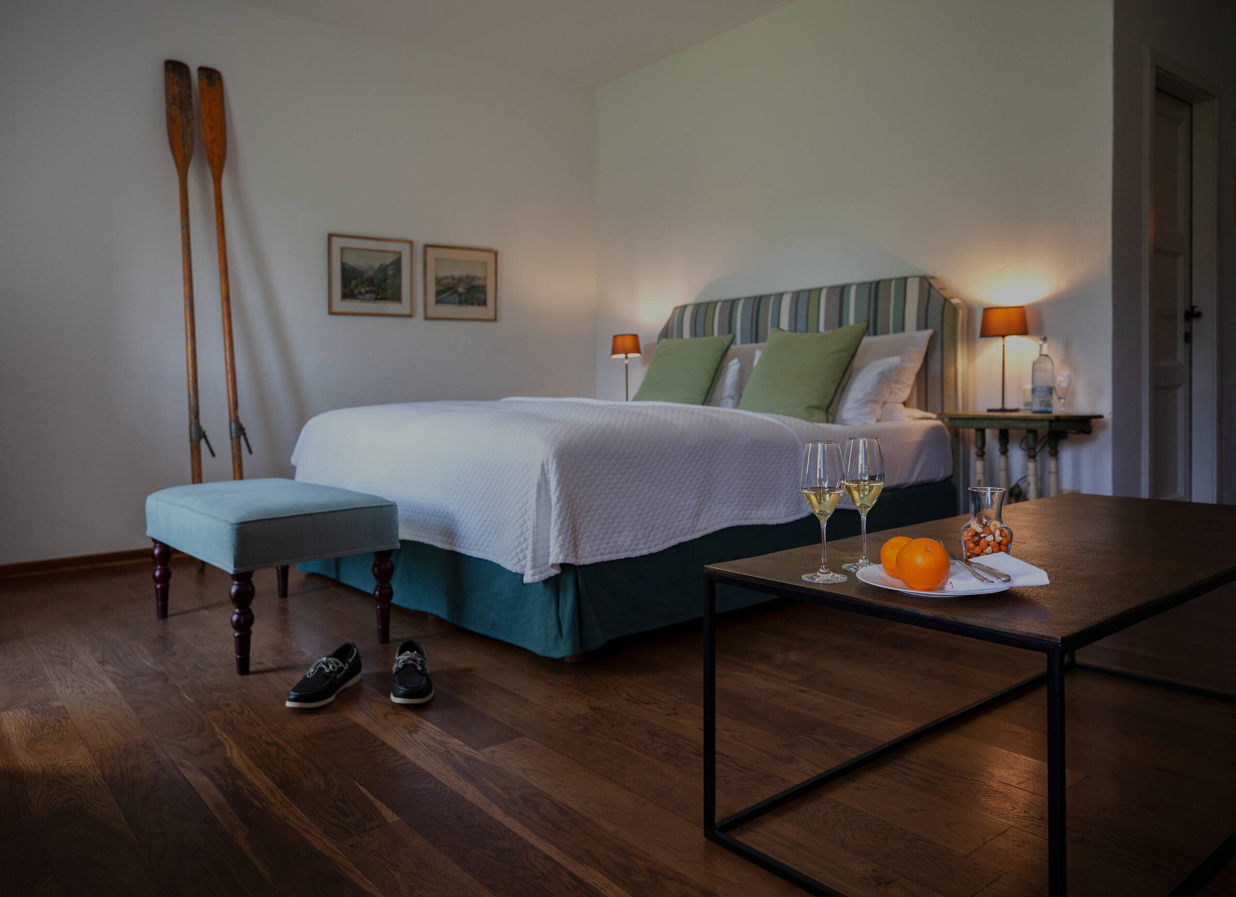 Ein helles Hotelzimmer mit einem großen Bett, grünen Kissen, Holzfußboden und zwei Weingläsern auf dem Tisch.