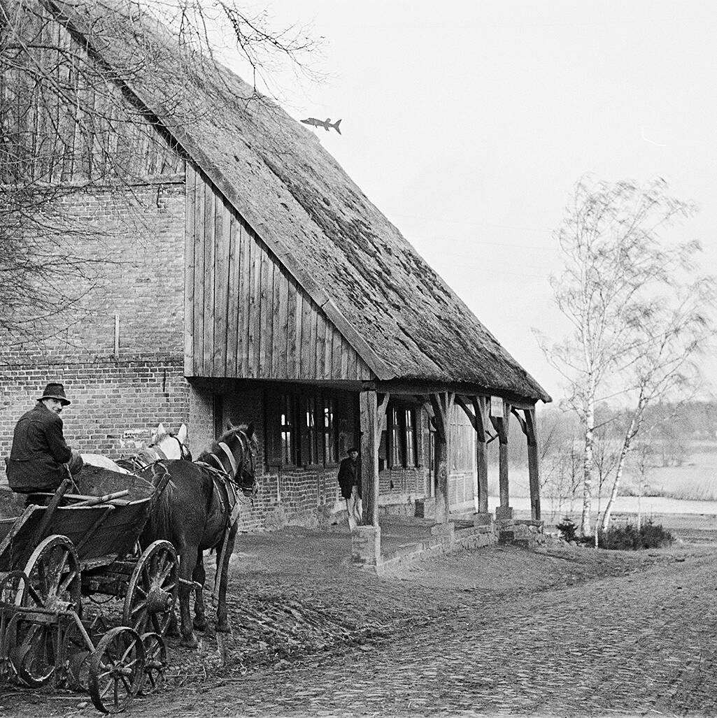 Eine Pferdekutsche vor einem Fachwerkhaus mit Strohdach, daneben steht eine Person am Haus, im Hintergrund Bäume und ein Feld.