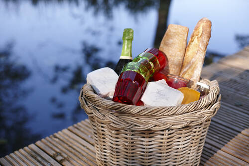 Ein geflochtener Picknickkorb mit frischem Baguette, Käse, Gläsern und Sektflasche auf einem Steg am Wasser.