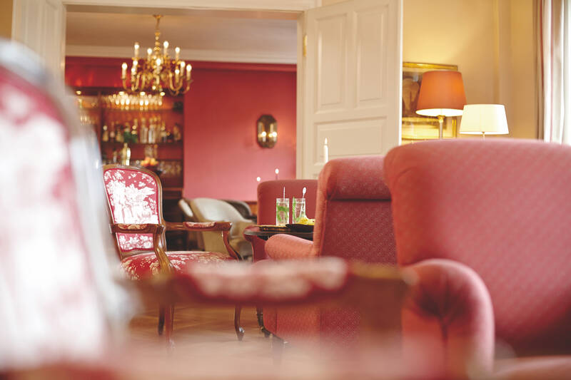 Ein Hotelzimmer mit roten Sesseln, einem Kronleuchter und einer offenen Tür zu einem Raum mit pinkfarbenen Wänden.