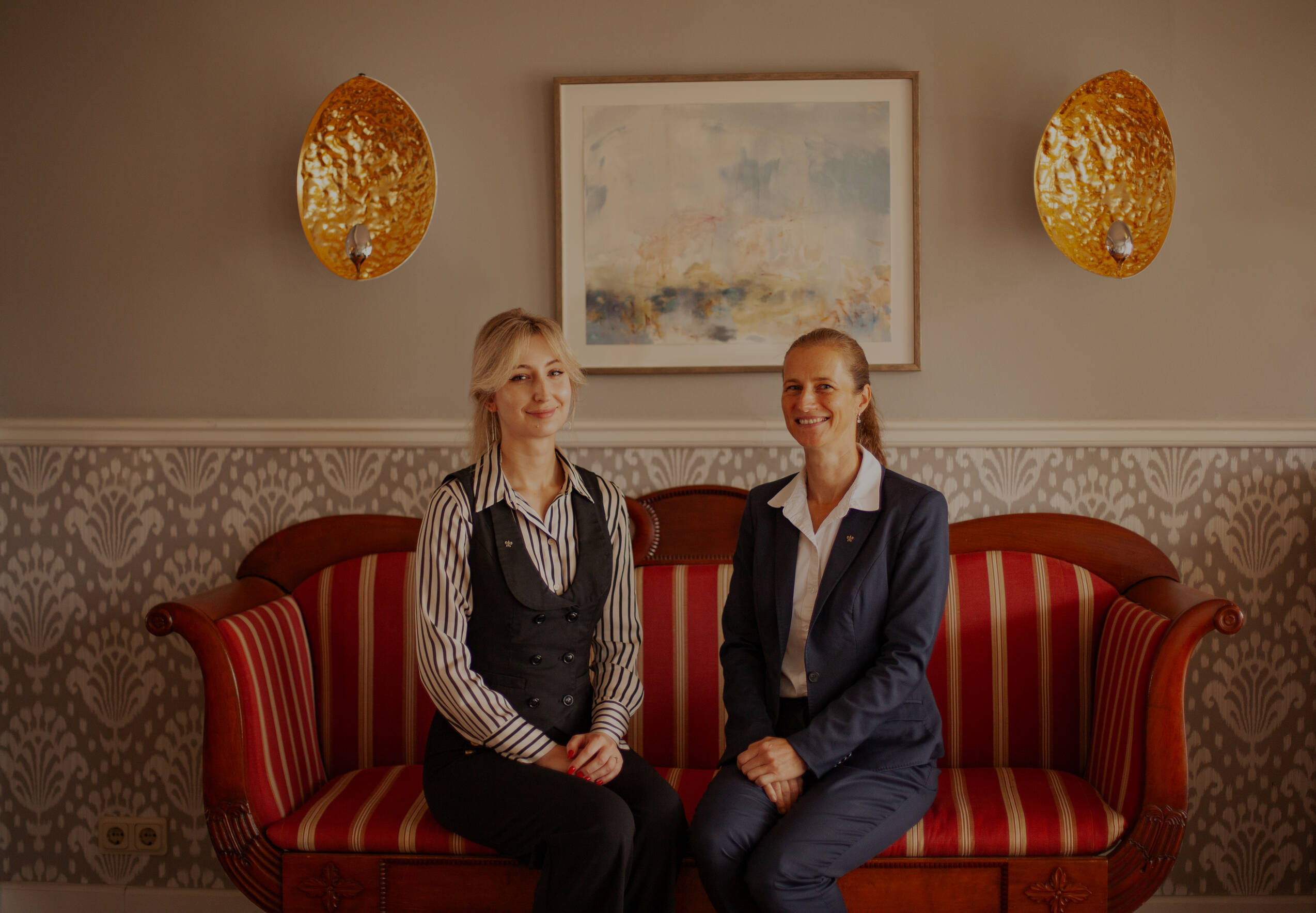 Zwei Frauen sitzen auf einem rot-weiß gestreiften Sofa vor einer Wand mit floralem Muster, darüber ein abstraktes Bild und zwei goldfarbene Wandlampen.