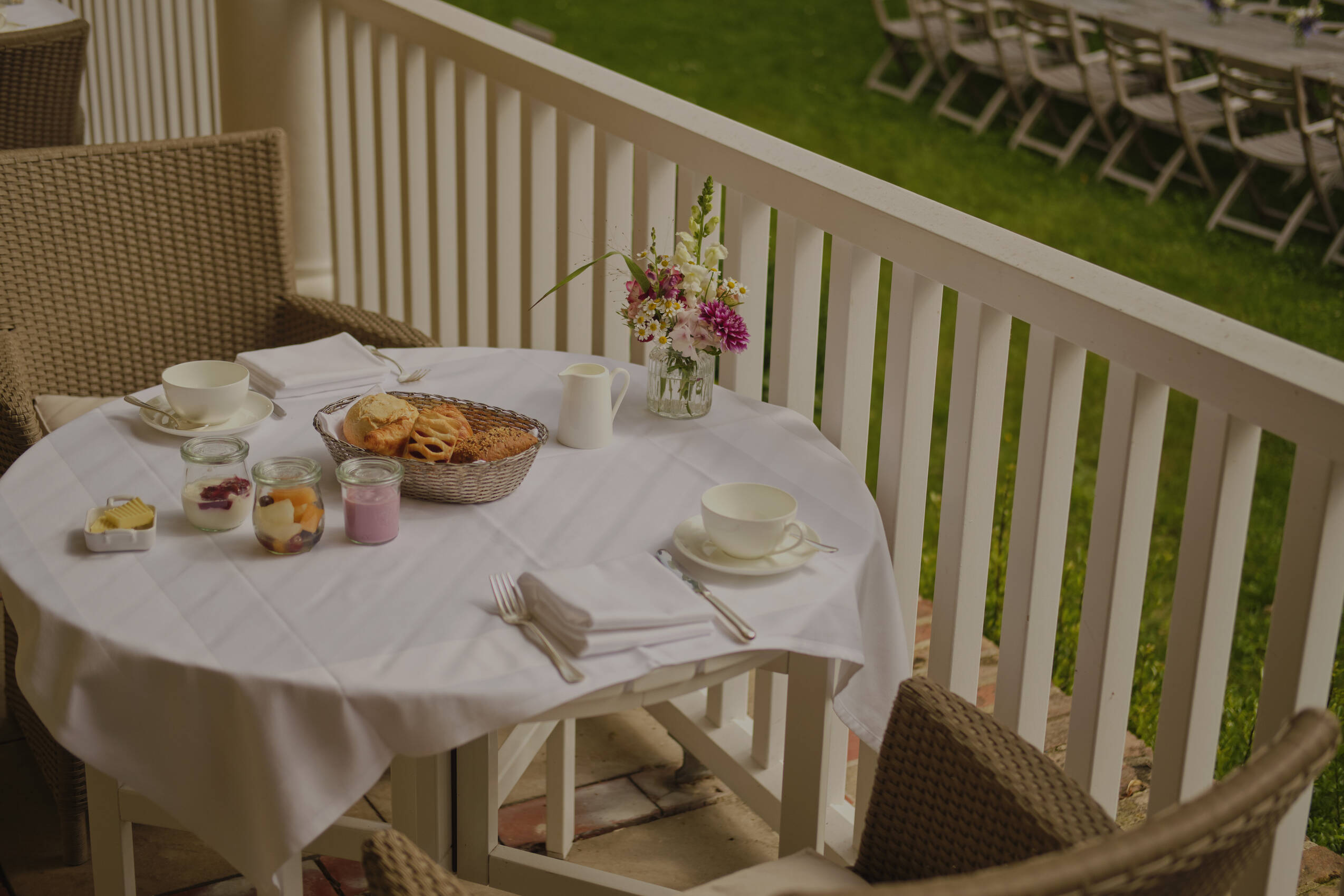 Ein runder Tisch mit einer weißen Tischdecke, gedeckt mit Tassen, Gläsern, Gebäck und einem Blumenstrauß, mit zwei Flechtstühlen auf einer Terrasse.
