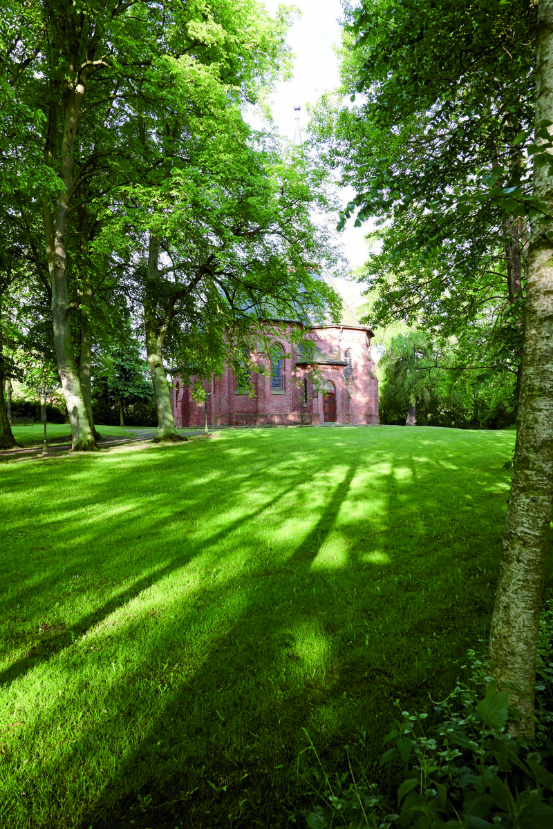 Eine rote Backsteinkirche mit Spitzbogenfenstern, umgeben von hohen Bäumen und grünem Rasen, im Sonnenlicht neben Gutshaus Stolpe.