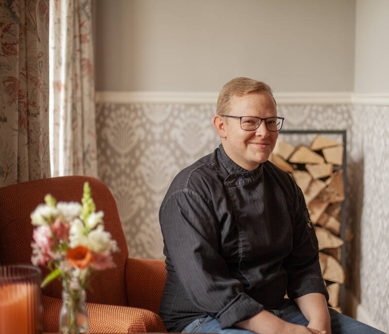 Eine Person in einer schwarzen Kochjacke sitzt auf einem orangefarbenen Sessel vor einer Wand mit einem floralen Muster und einem Holzstapel im Hintergrund.
