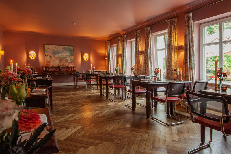 Ein gemütliches Restaurant nahe Anklam mit einem Holzboden, gedeckten Tischen, Stühlen mit Metallgestell und großen Fenstern mit gemusterten Vorhängen.