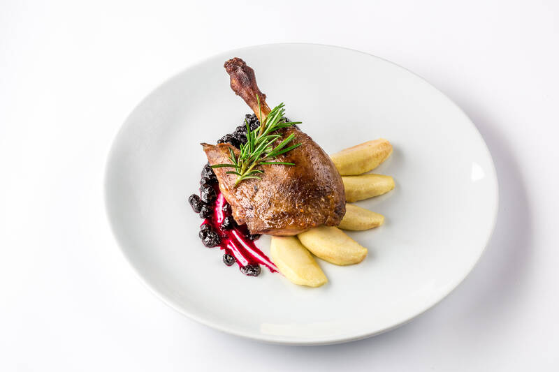 Eine gebratene Gänsekeule auf einem weißen Teller mit Kartoffeln, Blaubeeren und einem Zweig Rosmarin.