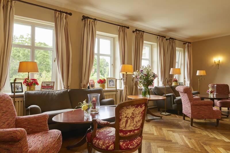 Eine elegante Hotellounge mit Sesseln, Sofas, Blumen und Lampen vor großen Fenstern mit hellen Vorhängen.