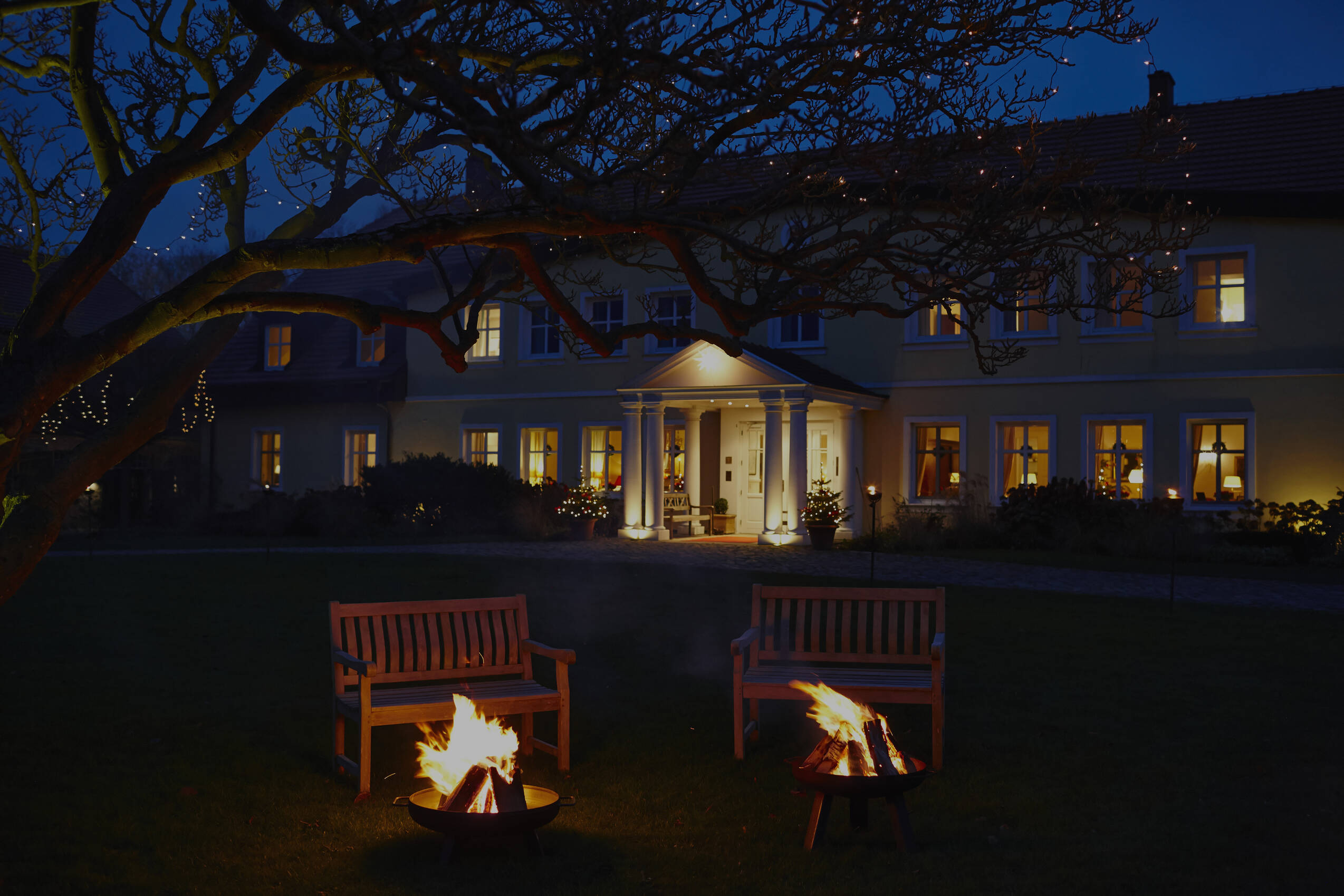 Ein beleuchtetes Herrenhaus am Abend mit zwei Feuerstellen und Holzbänken im Garten, umgeben von einem großen Baum.