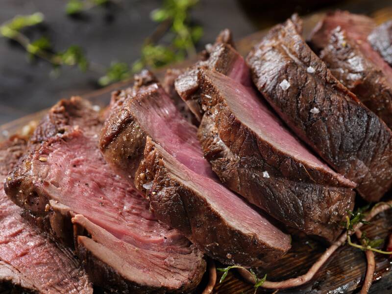 Mehrere Scheiben eines rosa gebratenen Rindersteaks liegen auf einem Holzbrett, garniert mit grobem Salz und Kräutern.