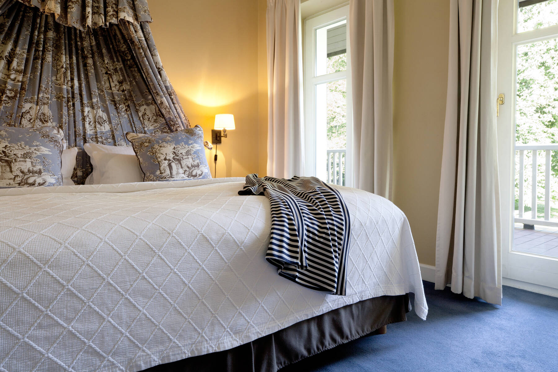 Ein Hotelzimmer mit einem großen Bett, einer weißen Steppdecke, einem gestreiften Pullover, einer Wandlampe und einem Balkon mit weißen Vorhängen.