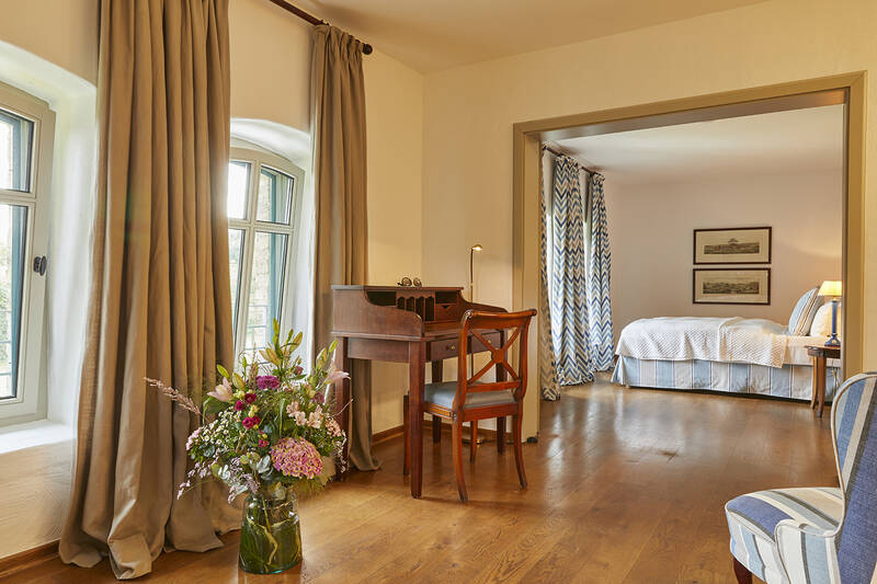 Ein helles Zimmer mit Holzboden, Fenster mit Vorhängen, einem Schreibtisch und Stuhl, einer Vase mit Blumen davor und einem angrenzenden Schlafzimmer.