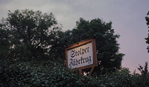 Ein Holzschild mit der Aufschrift 'Stolper Fährkrug' zwischen Bäumen bei Abendlicht.